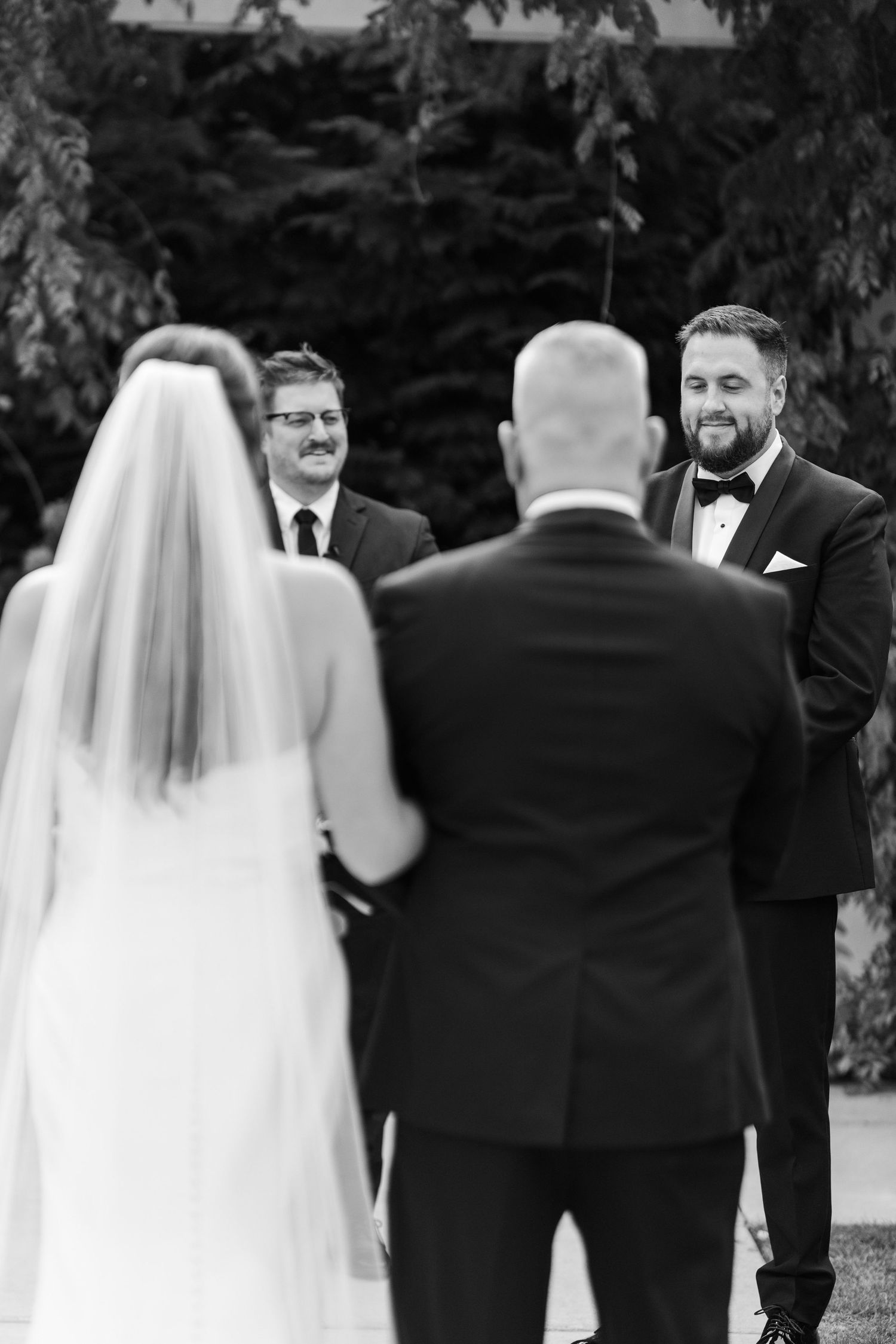 Groom smiling as bride walks down the aisle at outdoor wedding ceremony in black and white photo.