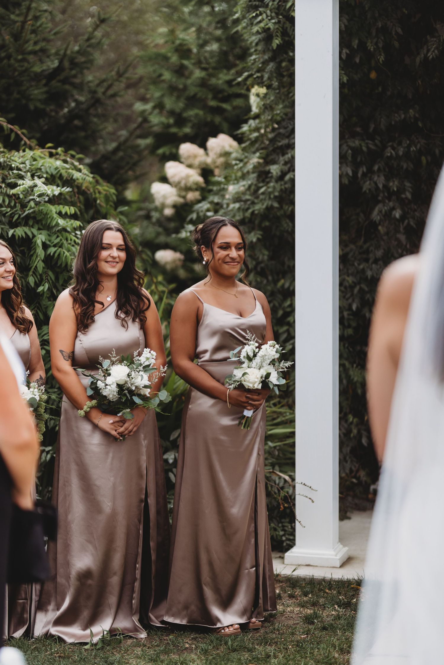 Bridesmaids in taupe satin dresses smile holding white bouquets at outdoor ceremony.