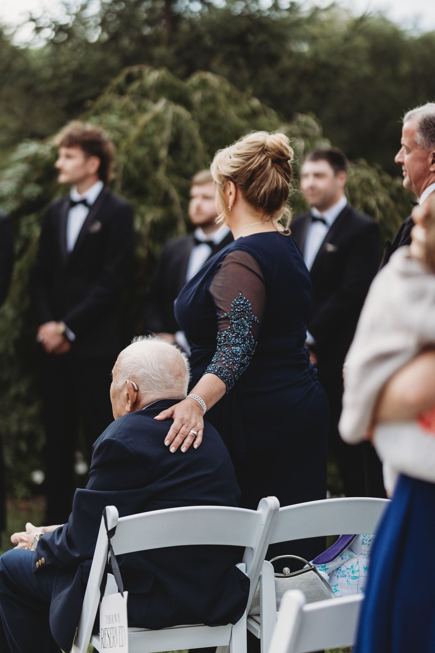 Woman in navy gown placing hand on elderly mans shoulder during outdoor wedding ceremony.