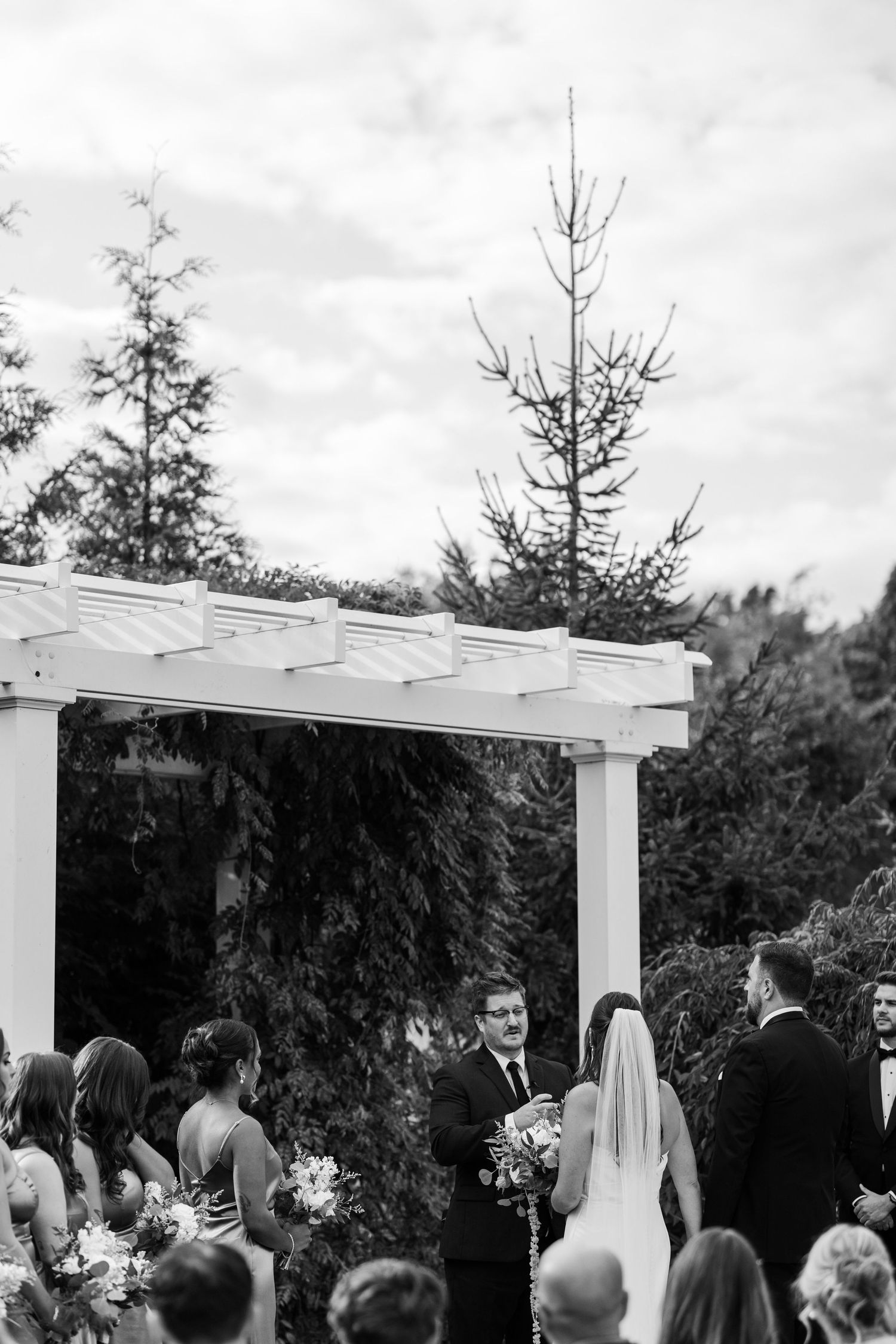 Black and white outdoor wedding ceremony under white pergola arbor surrounded by evergreen trees.