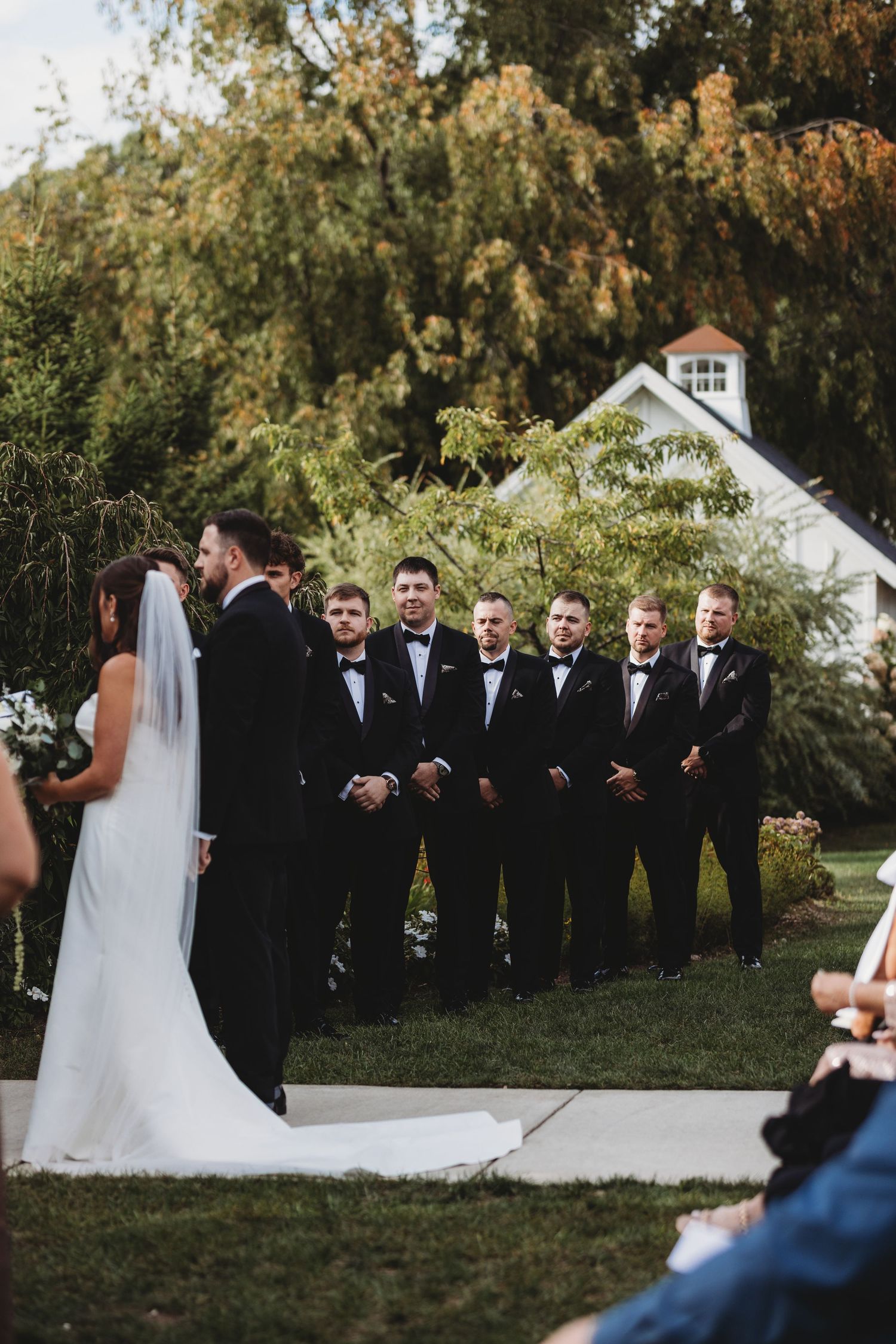 Groom and groomsmen in black tuxedos stand outdoors during wedding ceremony with bride in white gown and veil.