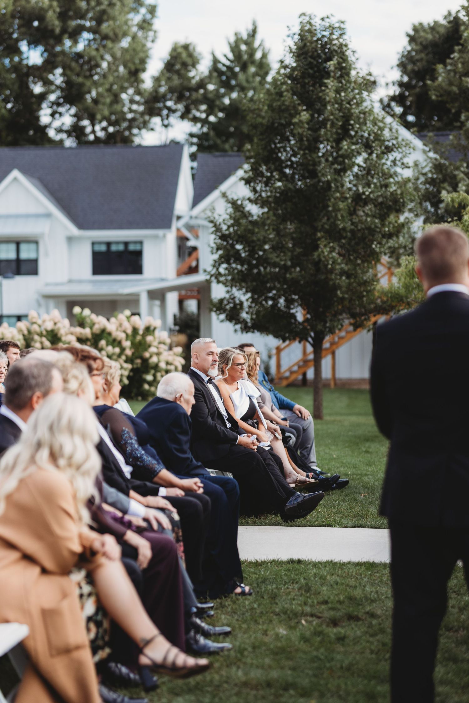 Wedding guests seated in outdoor ceremony chairs on green lawn watching groom await bride at rustic white farmhouse venue.