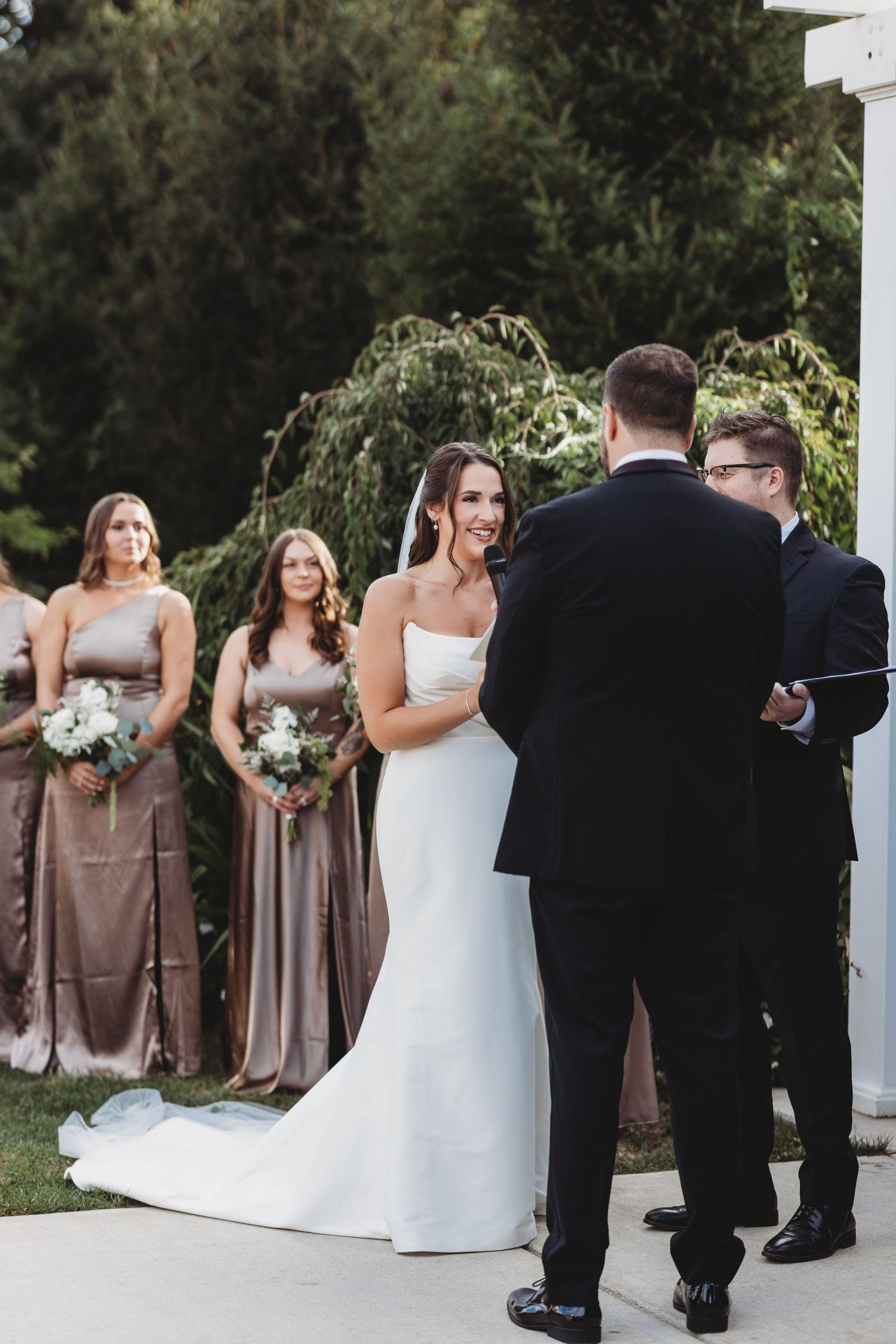 Bride smiles at groom during outdoor wedding ceremony with bridesmaids behind her.