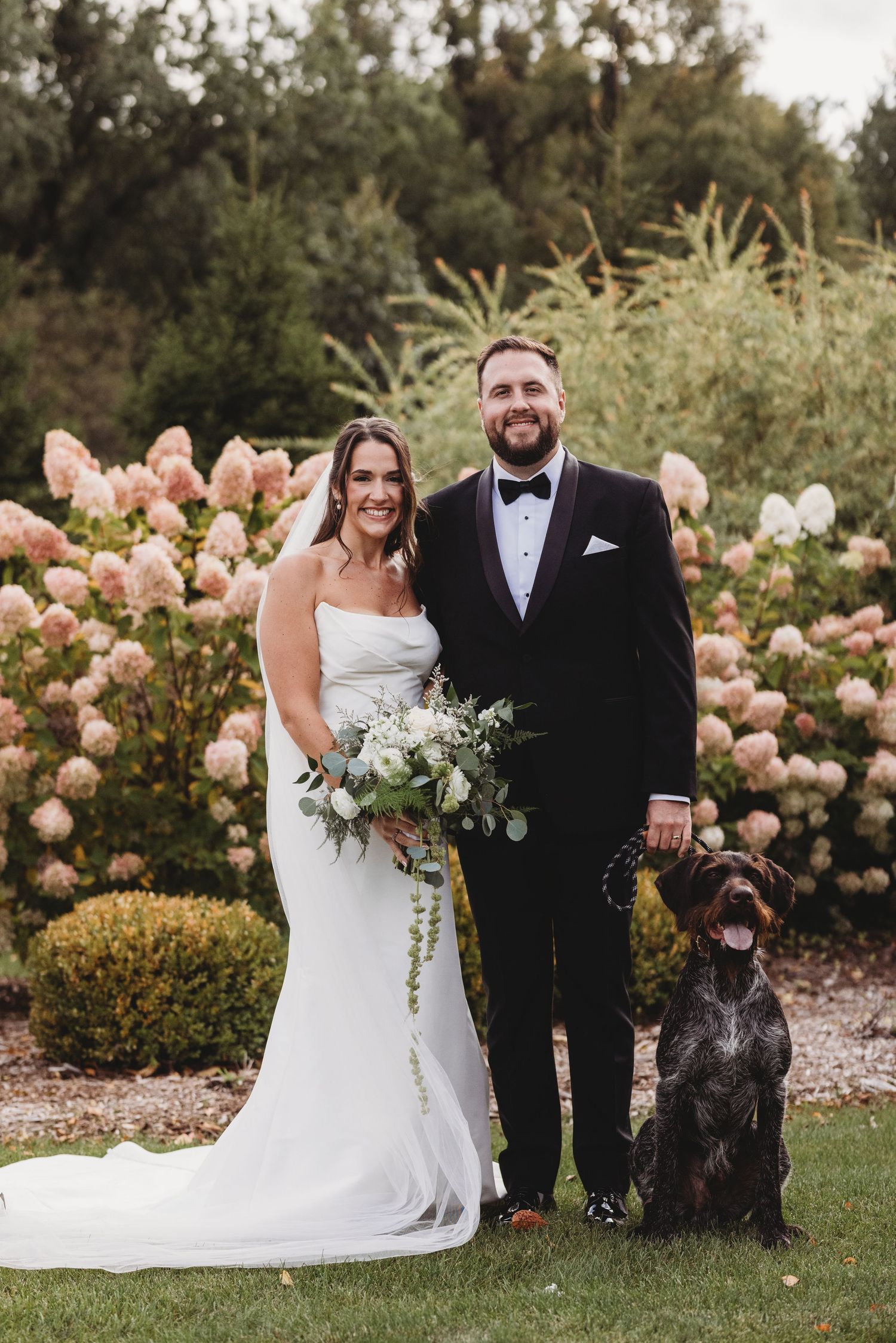 Bride and groom pose with their dog among pink hydrangeas in garden.