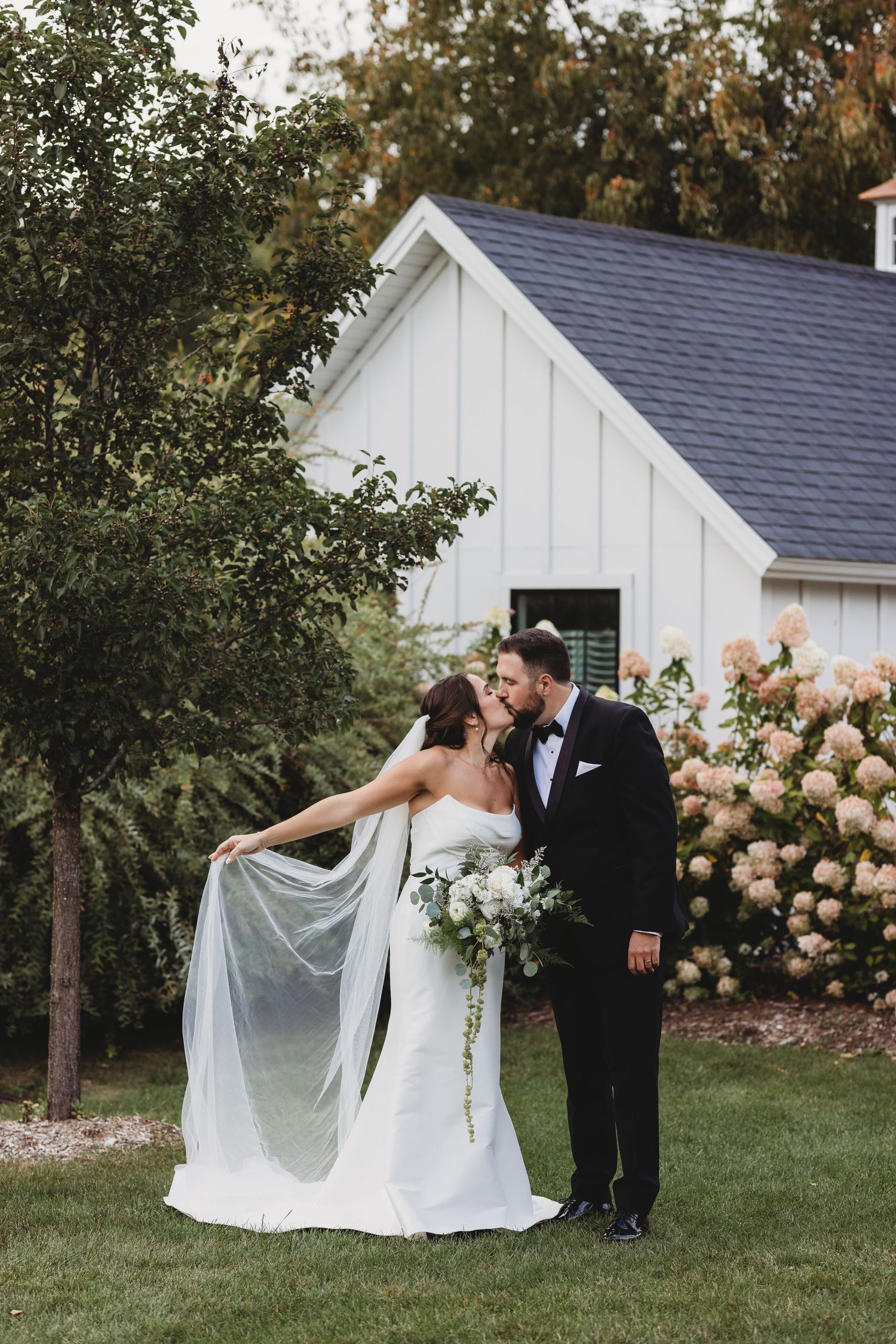 Bride and groom kissing near blooming hydrangeas and white barn at outdoor wedding venue.