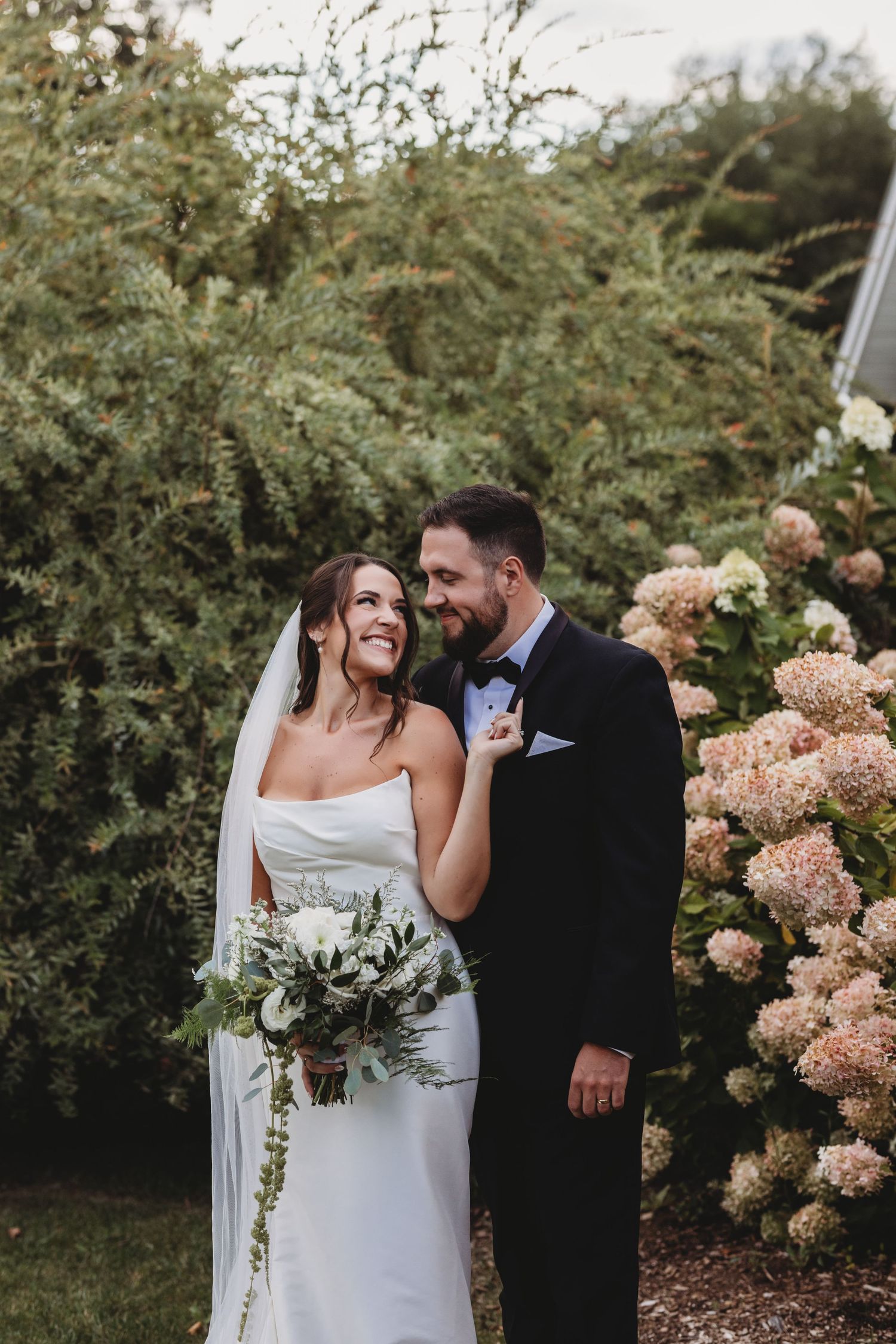 Bride and groom share romantic moment among lush greenery and pink hydrangeas.