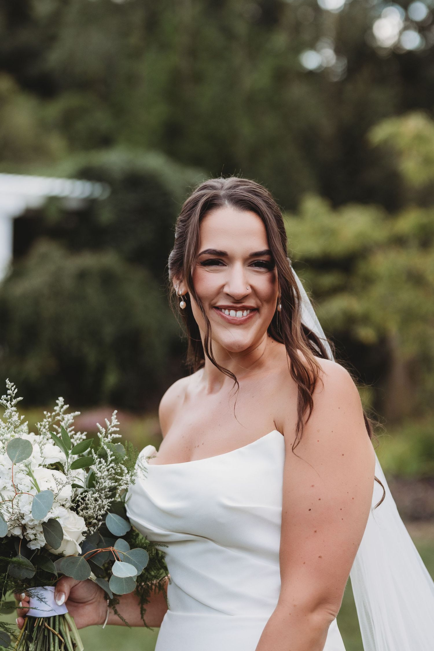 A smiling bride in a strapless white gown holds a lush bouquet of white flowers and eucalyptus outdoors.