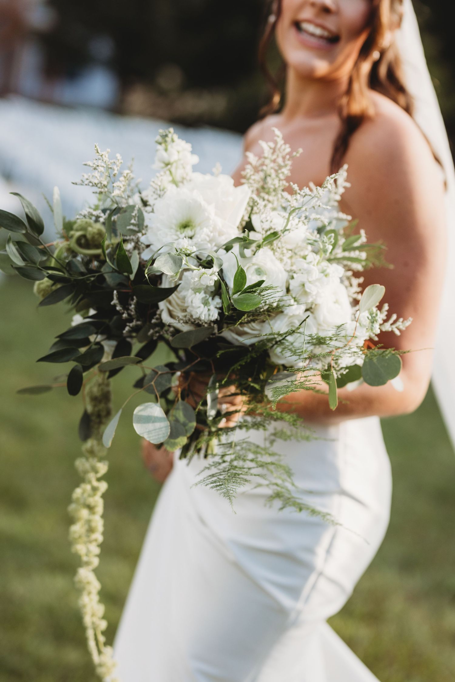 Smiling bride holding large white and green wedding bouquet with cascading greenery outdoors.