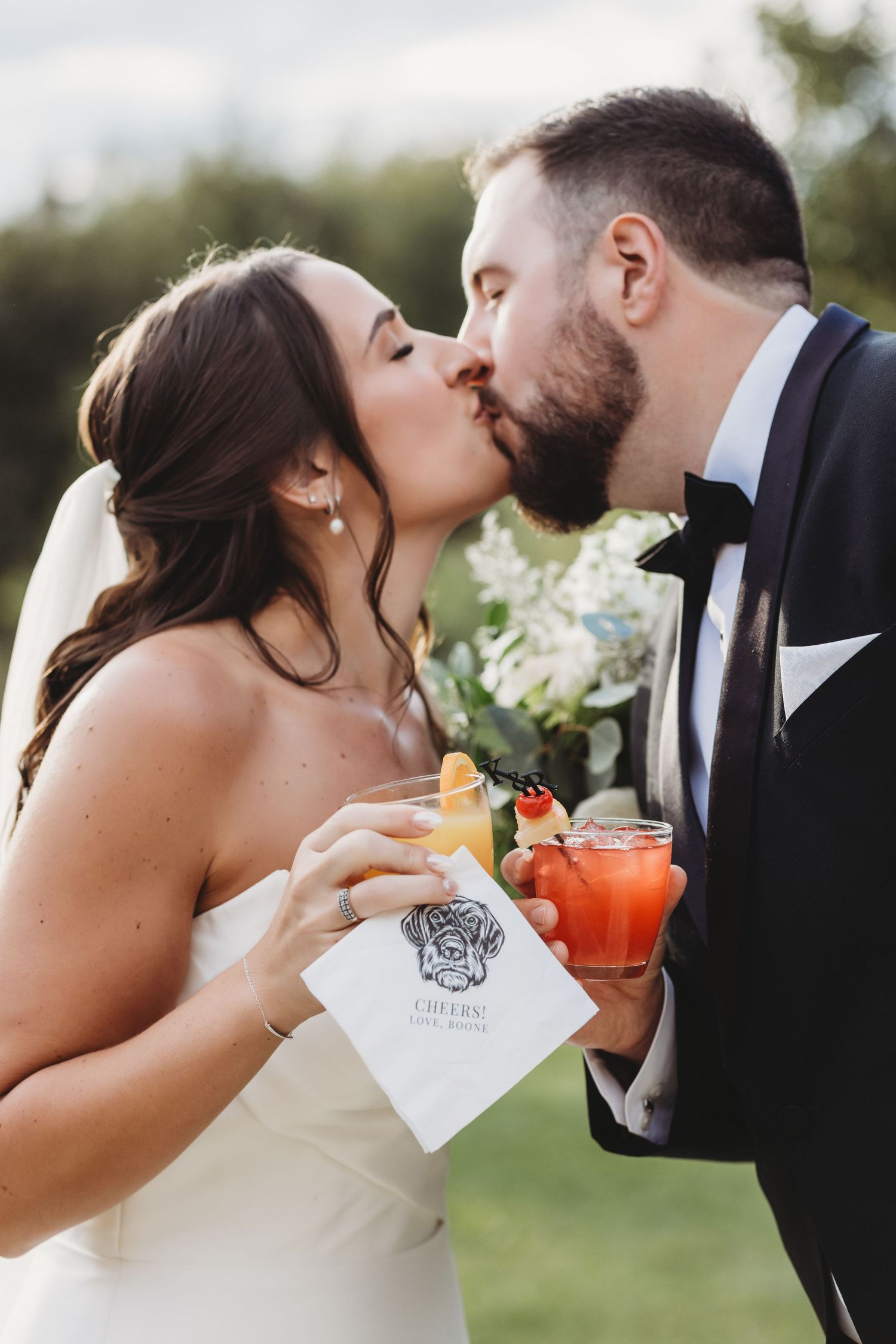 Bride and groom sharing first kiss while holding cocktails at outdoor wedding reception.