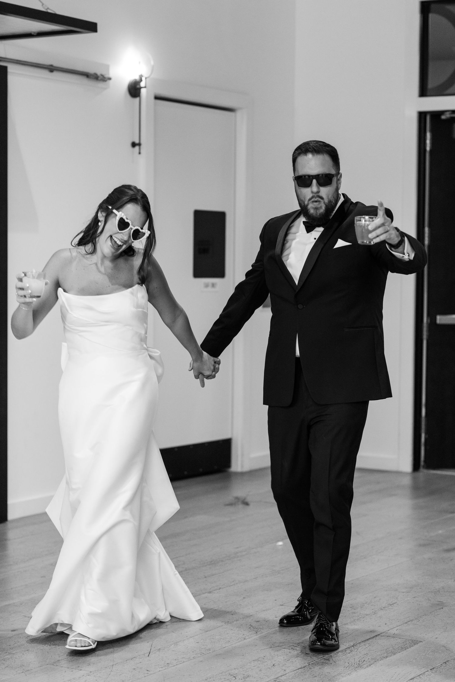 Black and white photo of a bride and groom holding hands, wearing sunglasses, and holding drinks at their wedding reception.