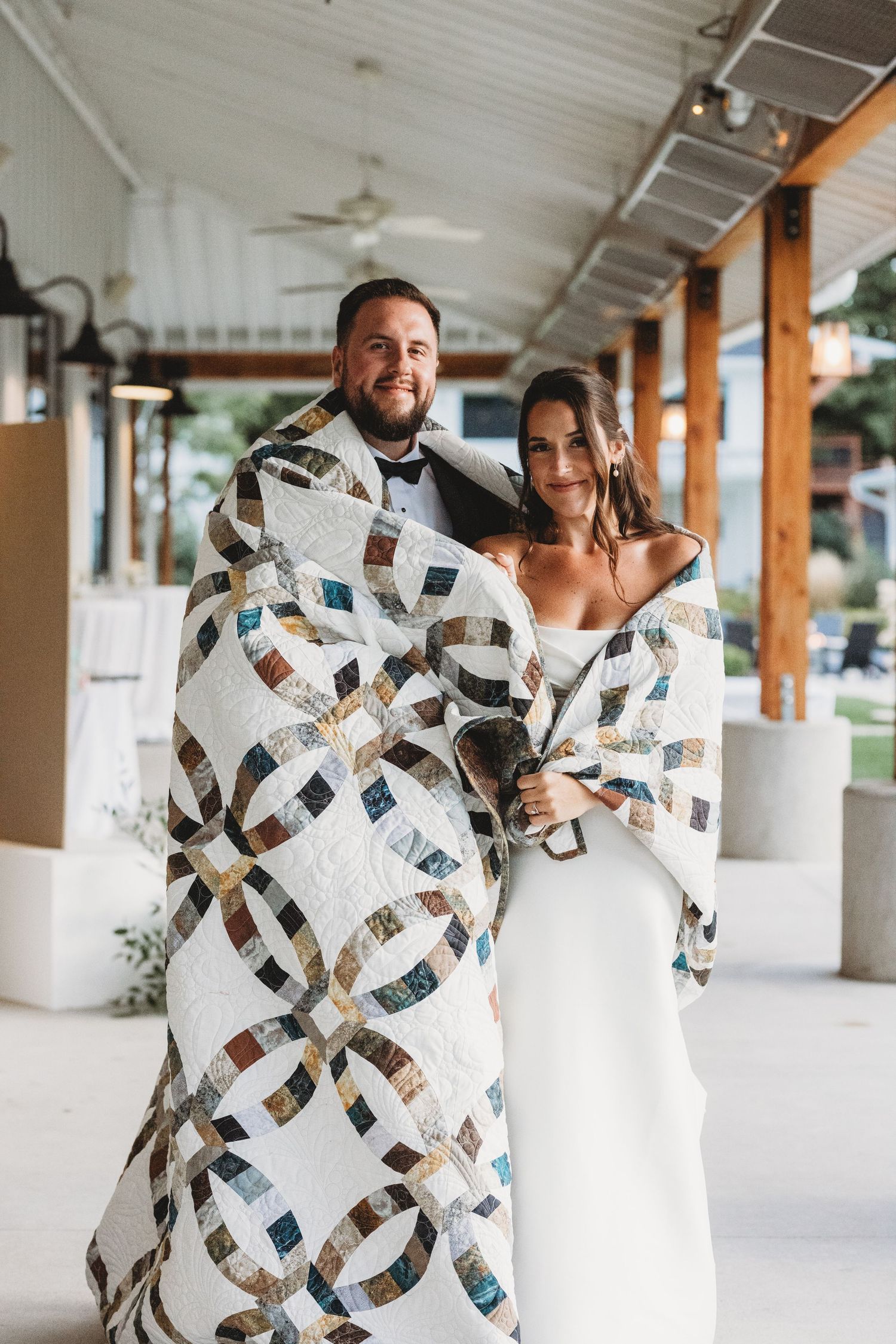 Bride and groom wrapped together in a colorful patchwork quilt at their wedding venue.