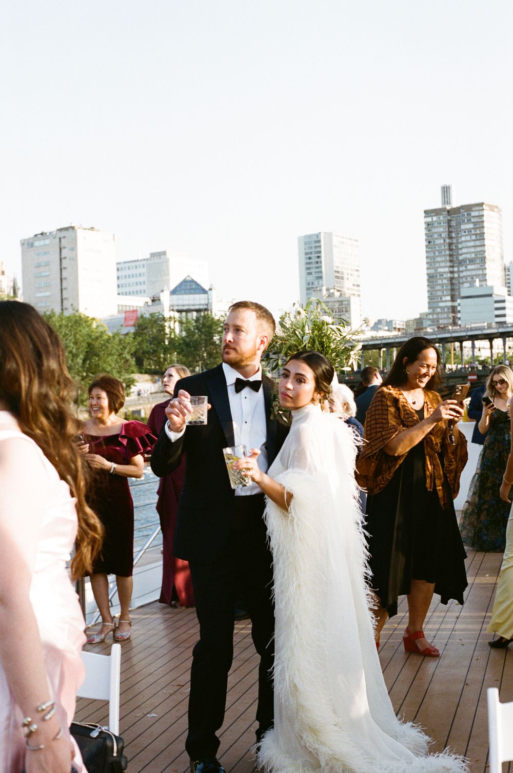 Bride in feather cape and groom in tuxedo walk through wedding reception on rooftop deck with city skyline backdrop. Paris wedding on boat