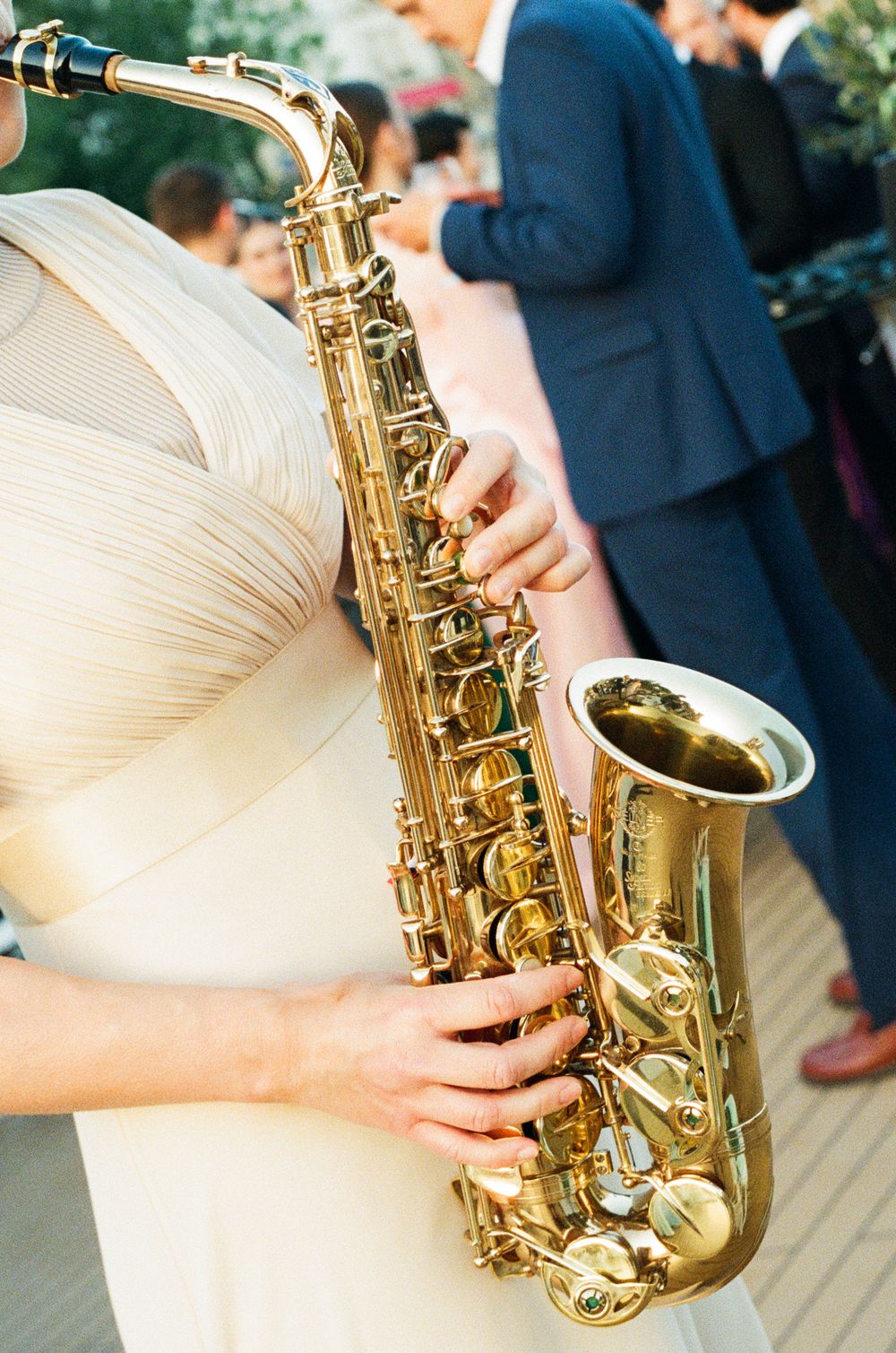 A musician in a cream dress plays a gleaming gold alto saxophone at an outdoor event.