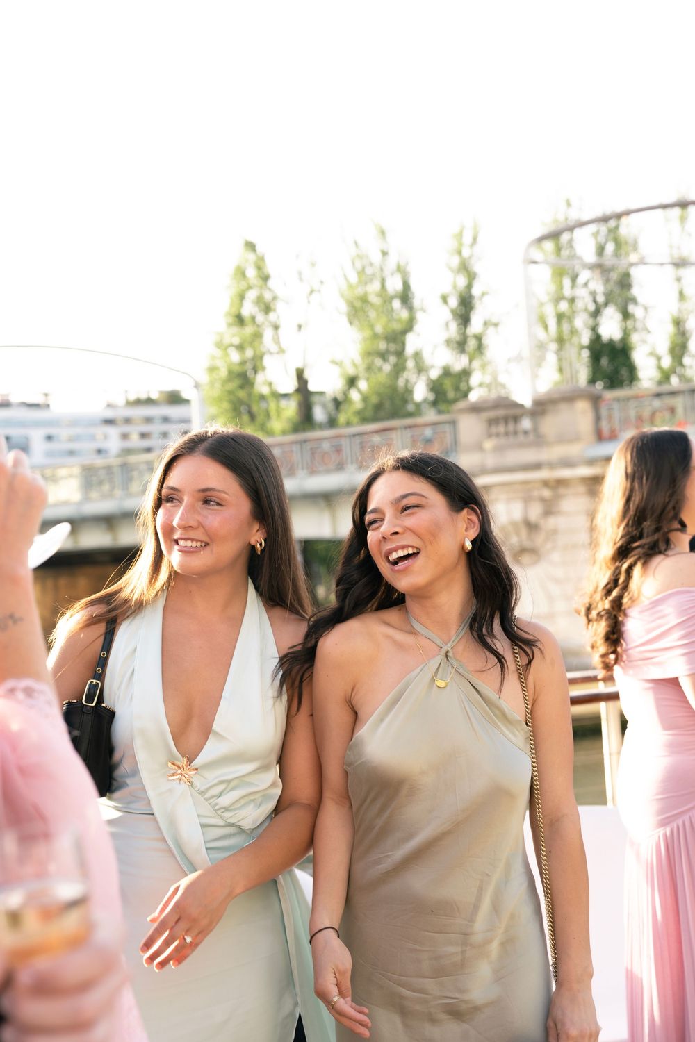 Two smiling women in elegant satin dresses at an outdoor event near a bridge.