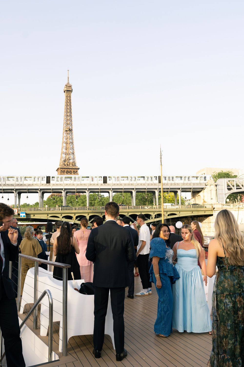 Elegantly dressed guests mingle on a boat deck with the Eiffel Tower and Bir-Hakeim bridge in Paris backdrop.