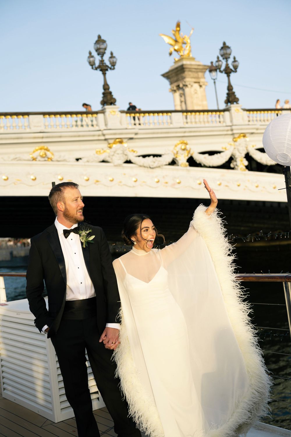 Bride in feathered cape and groom in tuxedo hold hands on Seine River boat, Pont Alexandre III bridge behind them. Best paris wedding