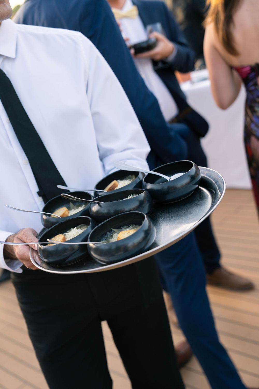 Waiter in white shirt carrying tray of black bowls with appetizers at outdoor catered event.