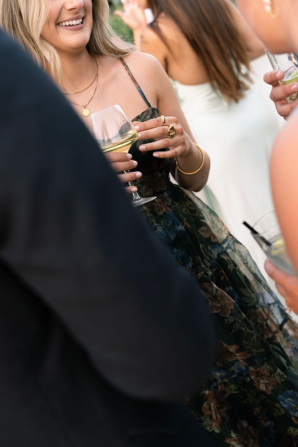 Guests socializing at an outdoor event, holding wine glasses, wearing elegant attire including floral skirt and gold jewelry. film wedding