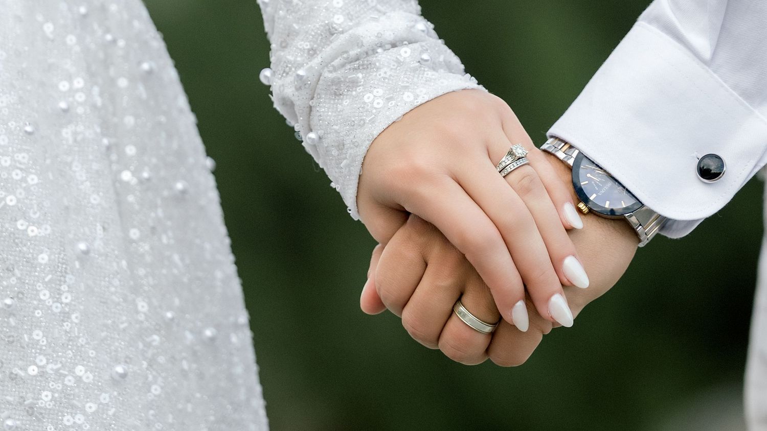 Newlywed couple holding hands, bride wearing diamond ring and white lace gown, groom in suit with cufflinks.