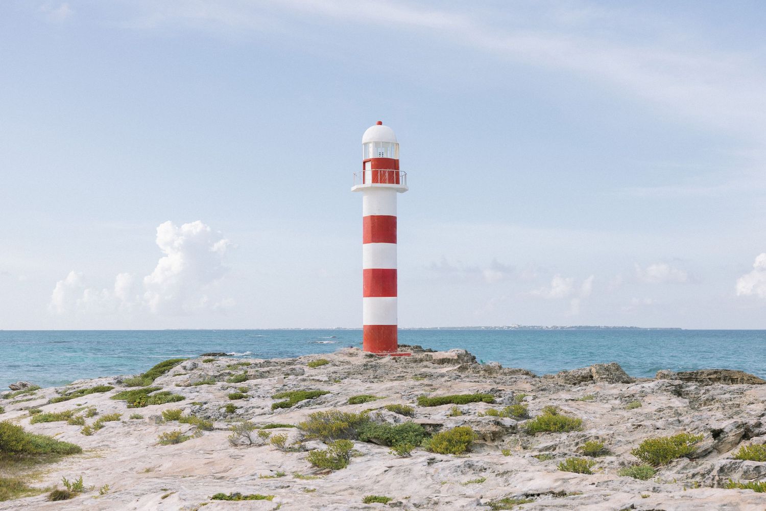 Red and white striped lighthouse standing on rocky coastal shoreline under partly cloudy sky.