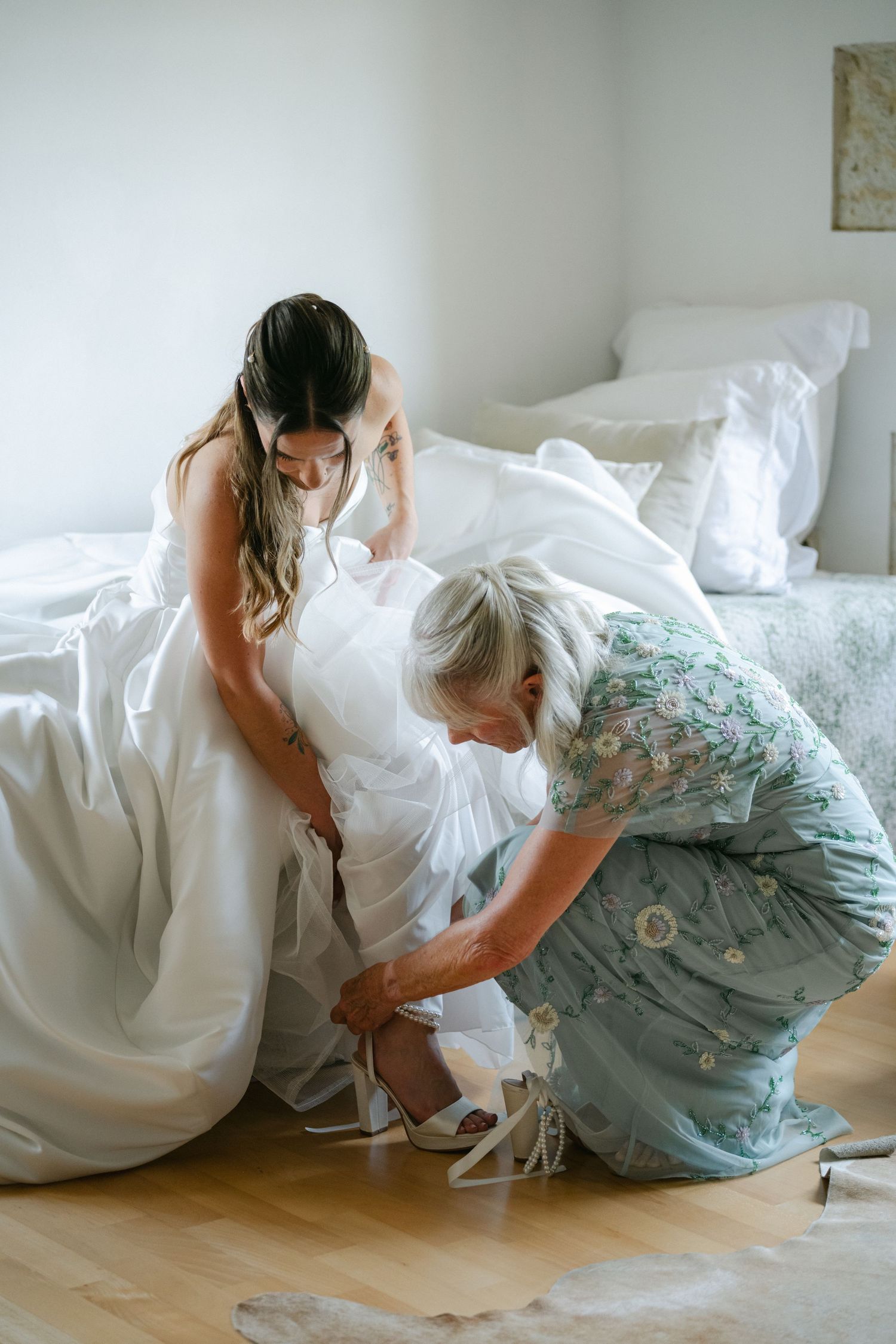 A bride in a white gown sits on a bed while a woman in a floral dress helps lace up her wedding shoes.