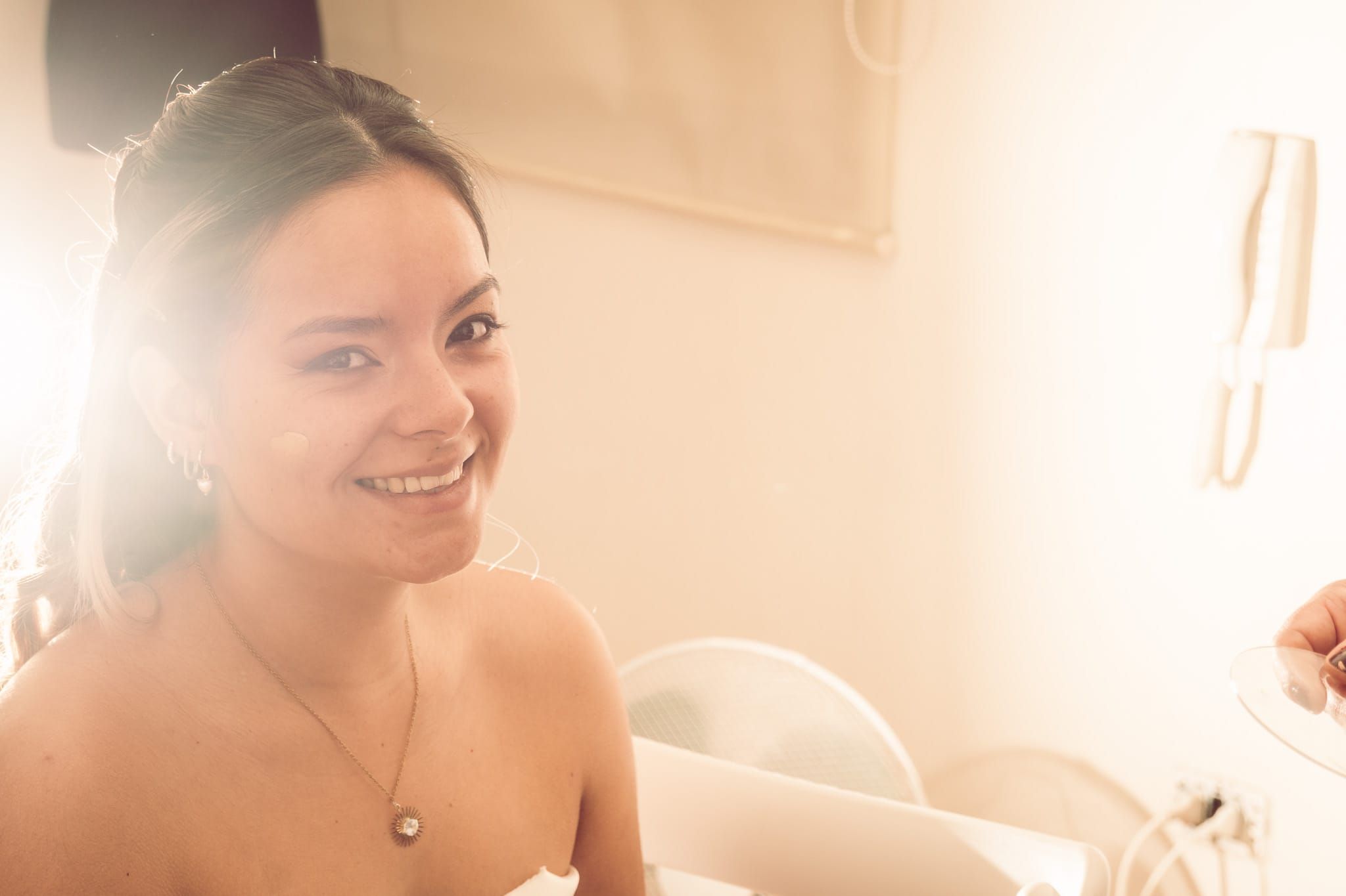 Smiling bride in white dress with necklace, glowing in warm sunlight during wedding preparations.