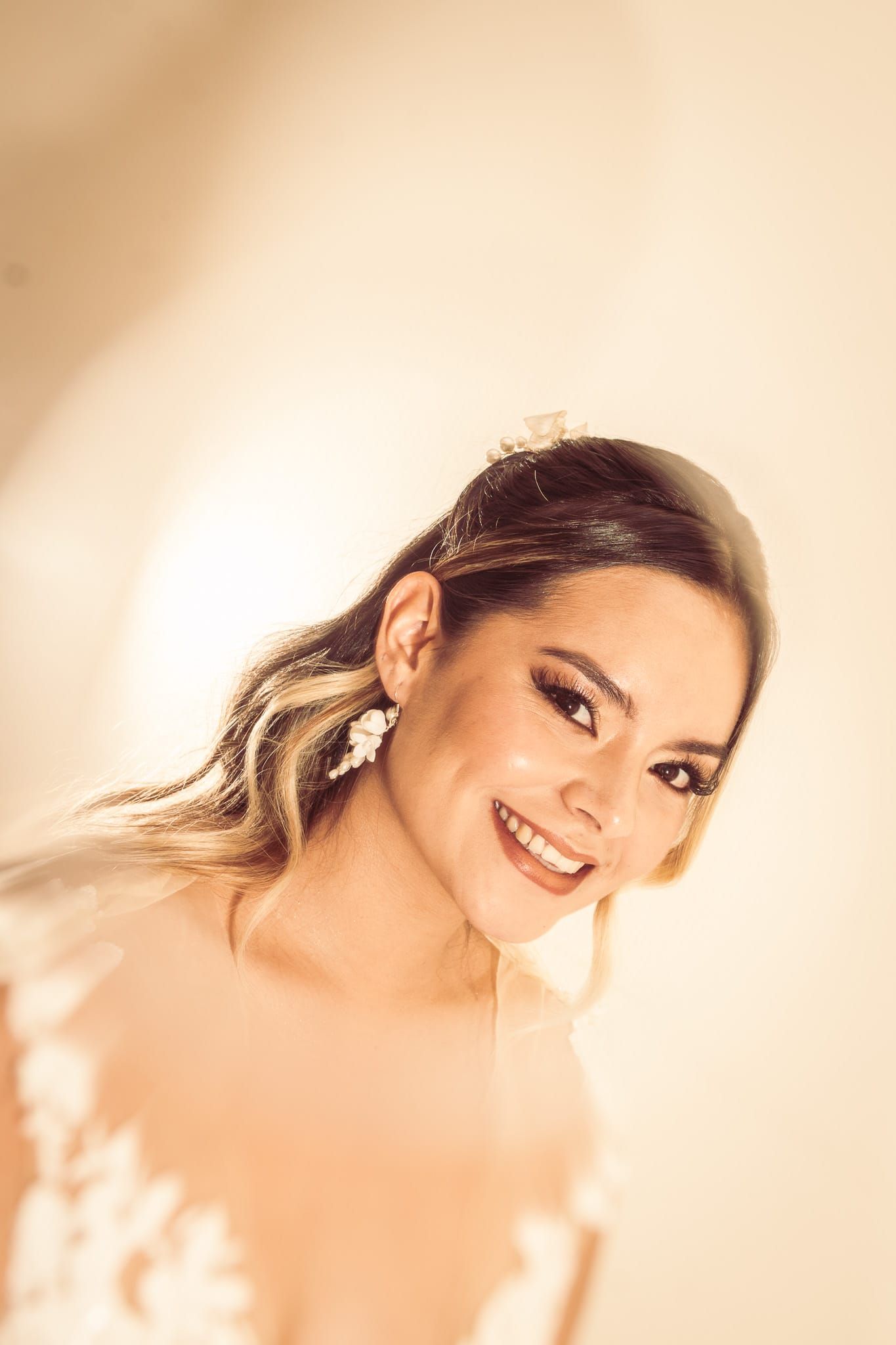 Beautiful bride smiling in warm light, wearing floral earrings and a lace wedding dress with elegant hair and makeup.