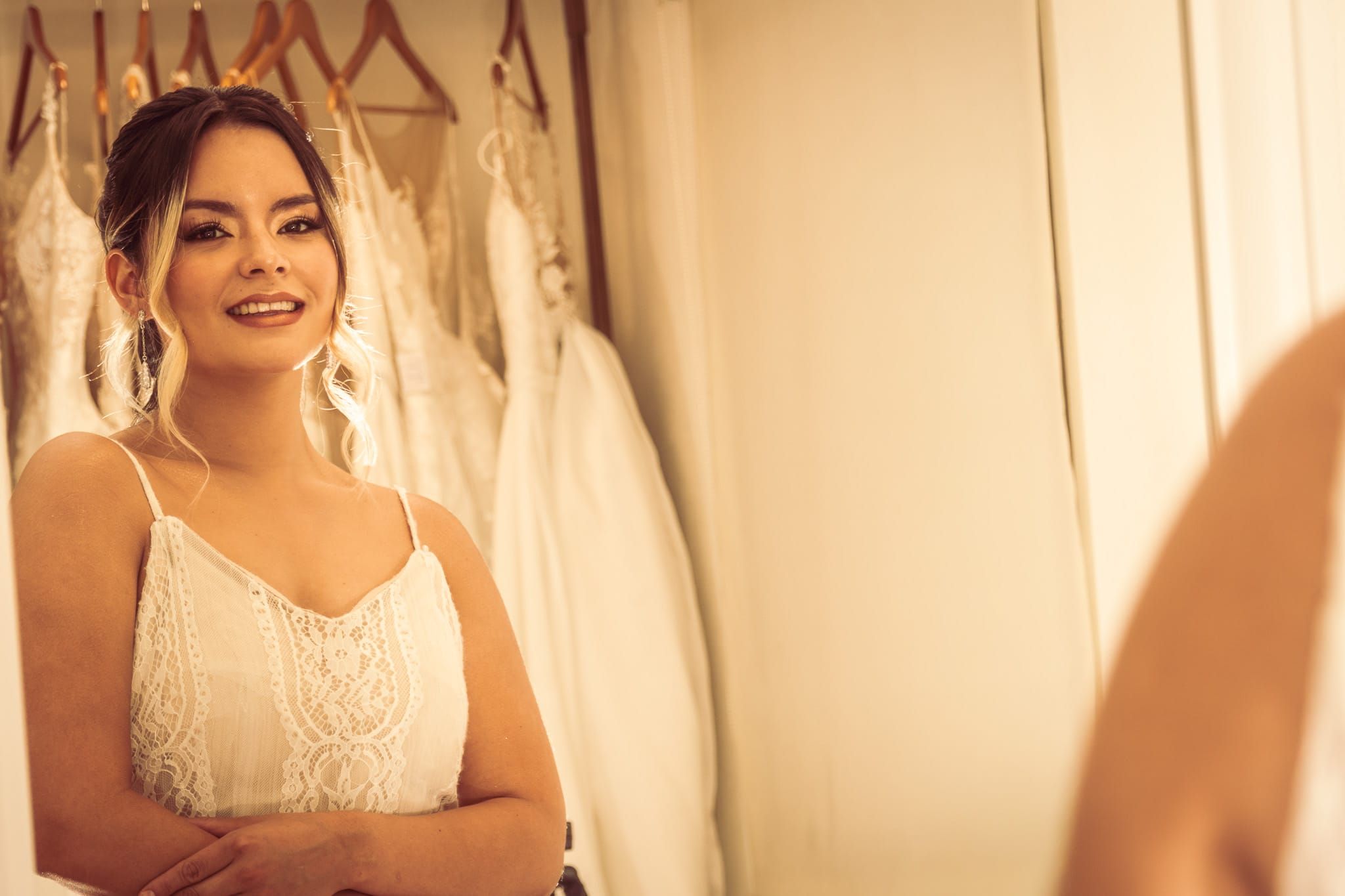 Smiling bride in white dress with arms crossed standing in bridal boutique with gowns on rack behind her.