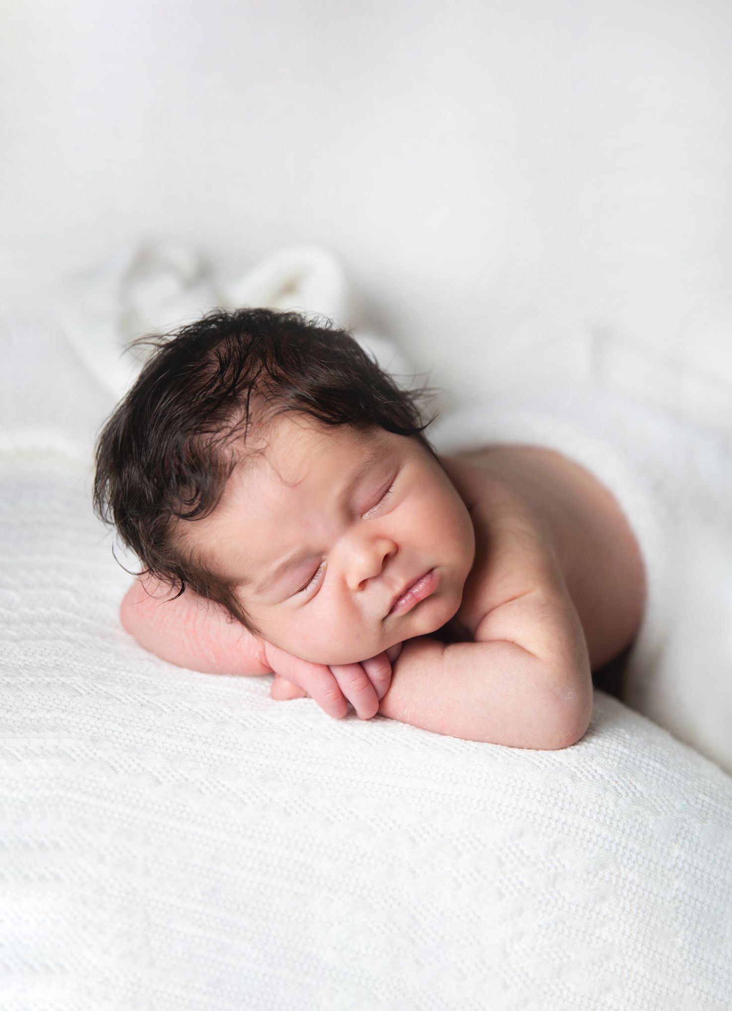 Peaceful newborn baby with dark curly hair sleeping on a white blanket in a classic froggy pose. During a newborn session in Bromley