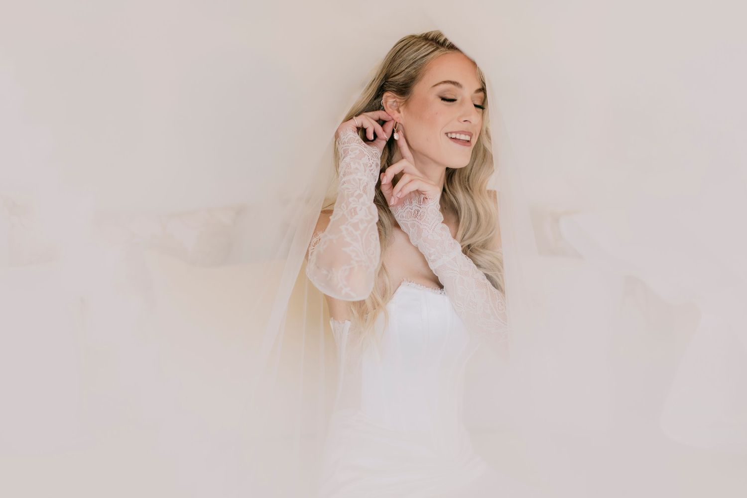 Smiling blonde bride in a white gown adjusting her earring, wearing lace gloves against a soft neutral background.