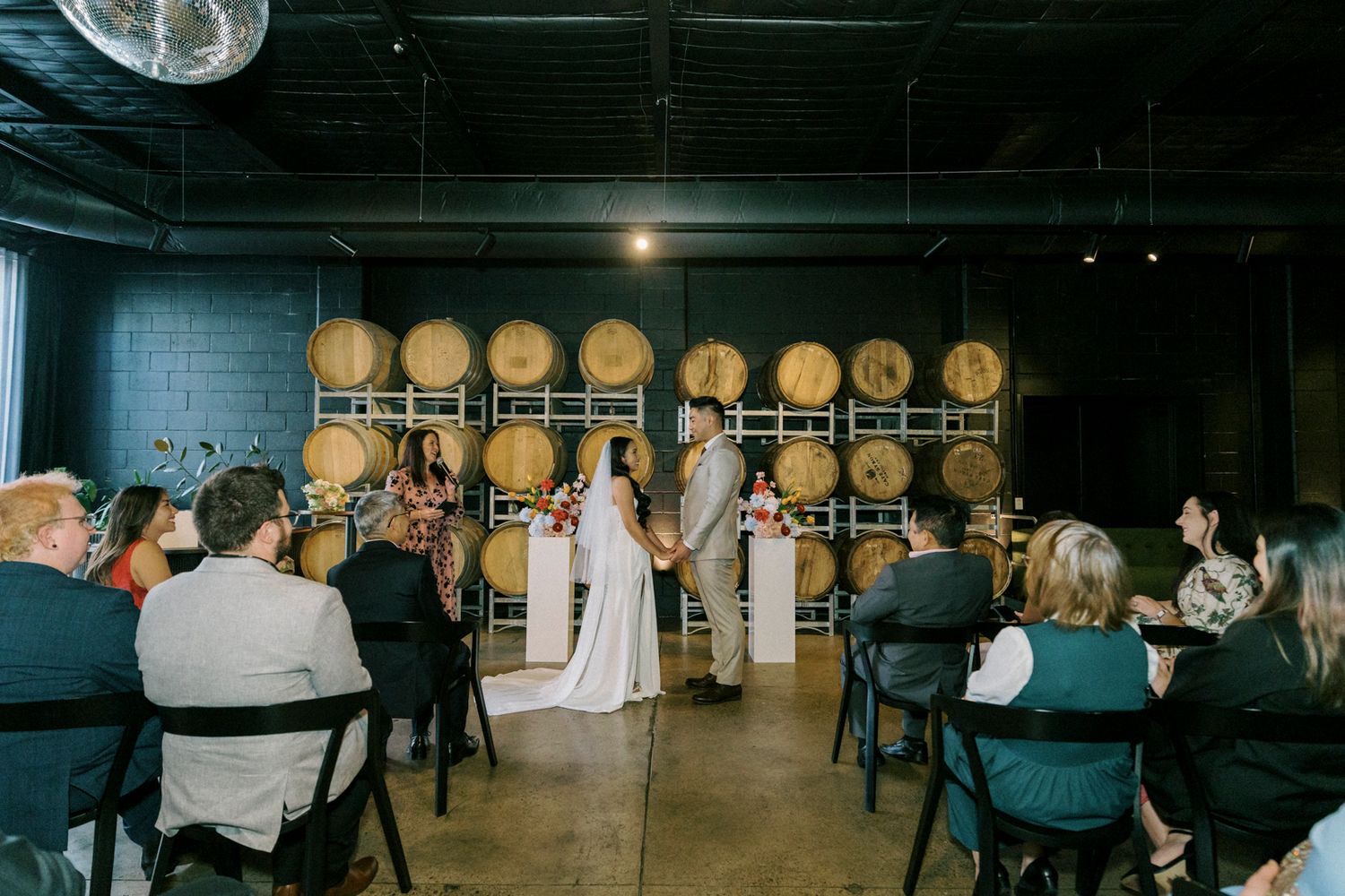 Wedding ceremony in a winery with barrel backdrop, bride and groom at altar surrounded by seated guests.
