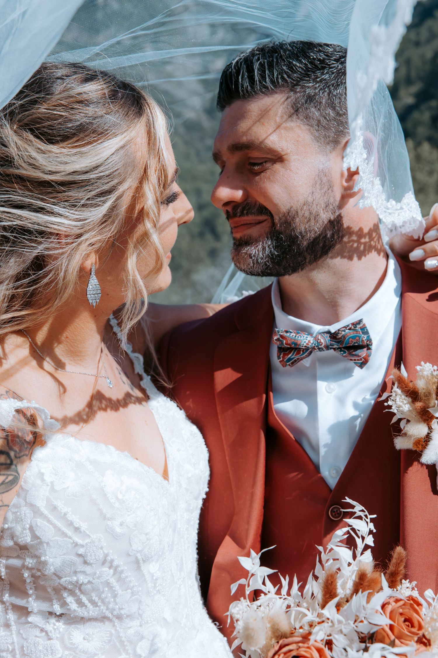 A smiling bride and groom share a romantic moment under a veil, with the groom in a rust-red suit and bow tie.