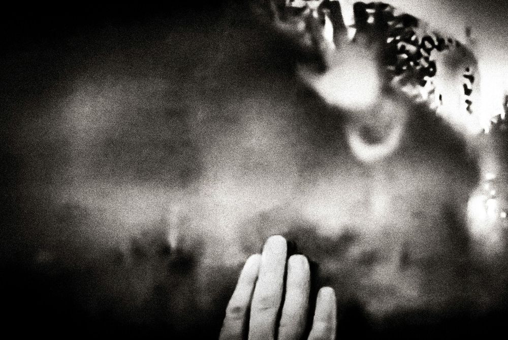 Black and white photo of a hand pressed against glass with blurred figures in the background.