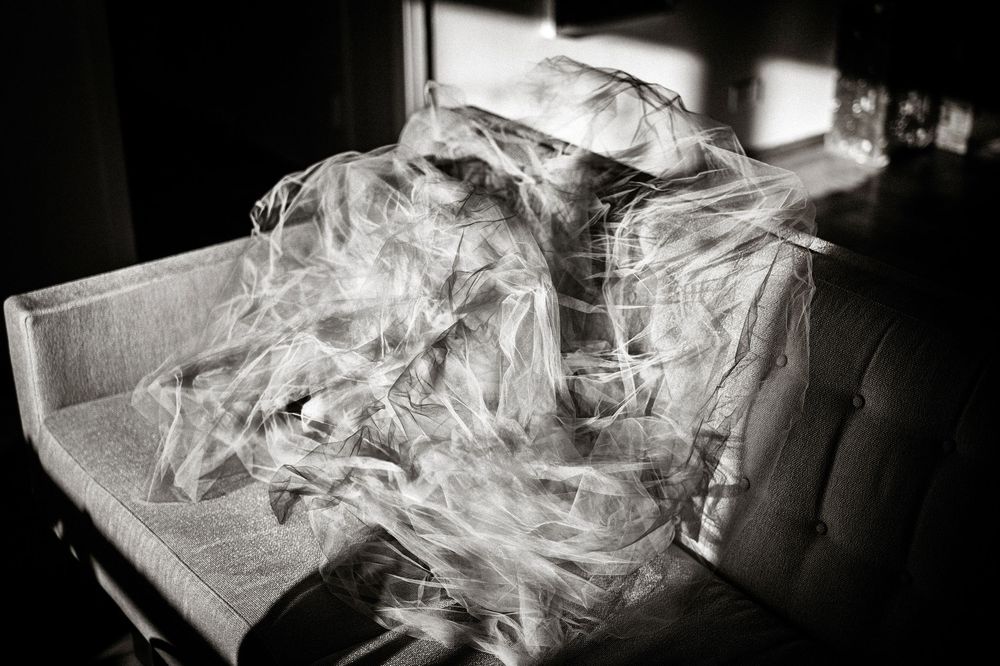 Black and white photo of a person wrapped in flowing white tulle fabric on a light sofa near a window.