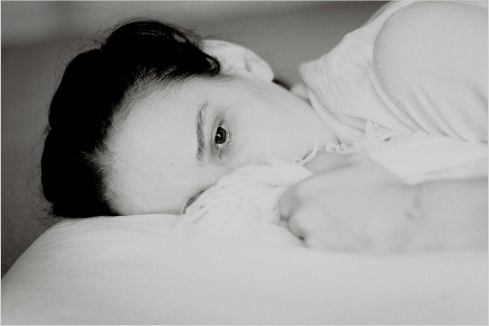 Black and white photo of a young woman lying in bed, resting her head on a pillow with a contemplative gaze.