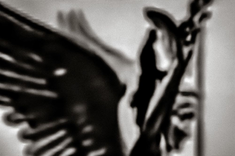 Black and white silhouette of a winged angel statue holding a staff against a dramatic dark background.