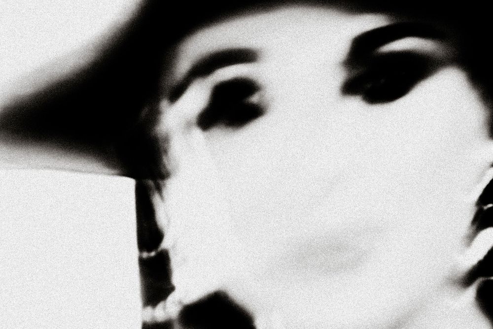 Black and white blurred portrait of a person wearing a wide-brim hat and pearl necklace.