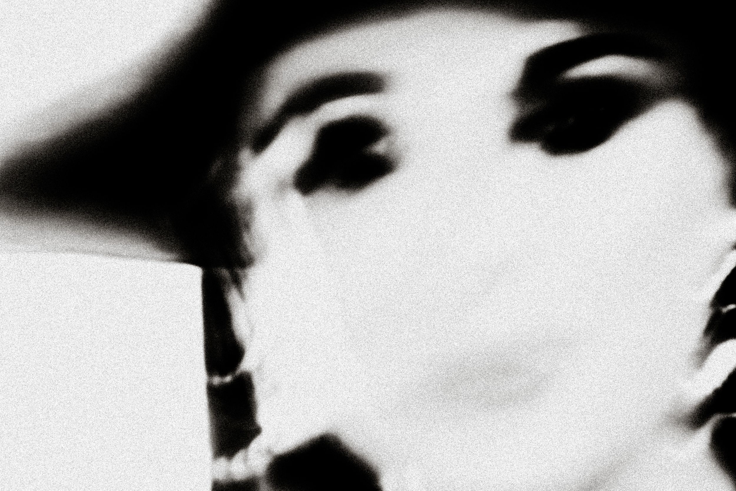 Black and white blurred portrait of a person wearing a wide-brim hat and pearl necklace.