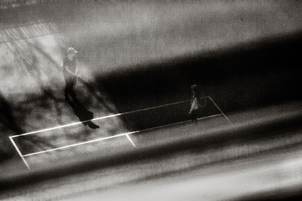 Two figures play tennis on a court in a dramatic black and white motion-blur photograph.