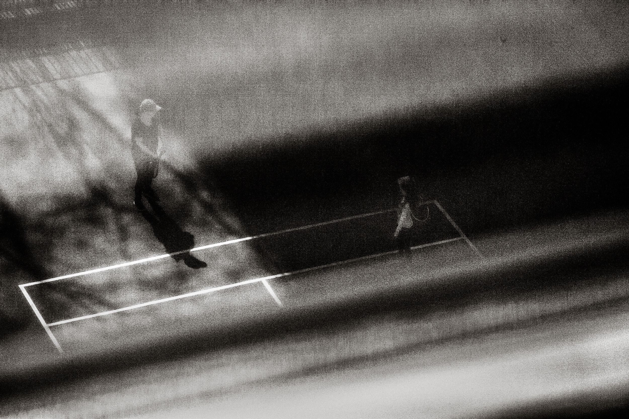 Two figures play tennis on a court in a dramatic black and white motion-blur photograph.