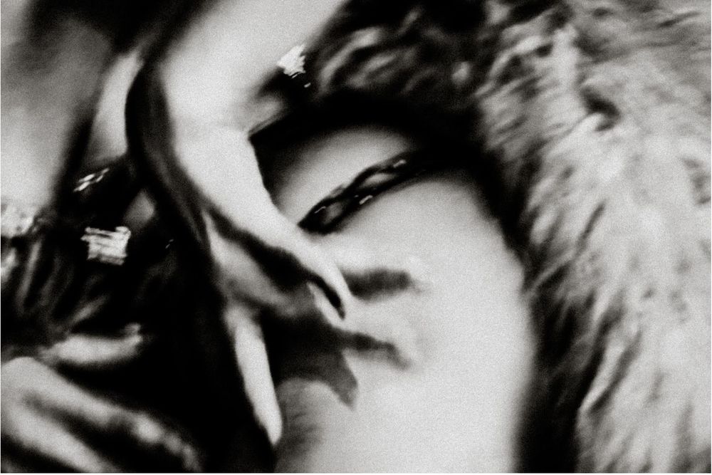 Black and white close-up photo of a woman with dramatic shadows, wearing fur, hand near face.