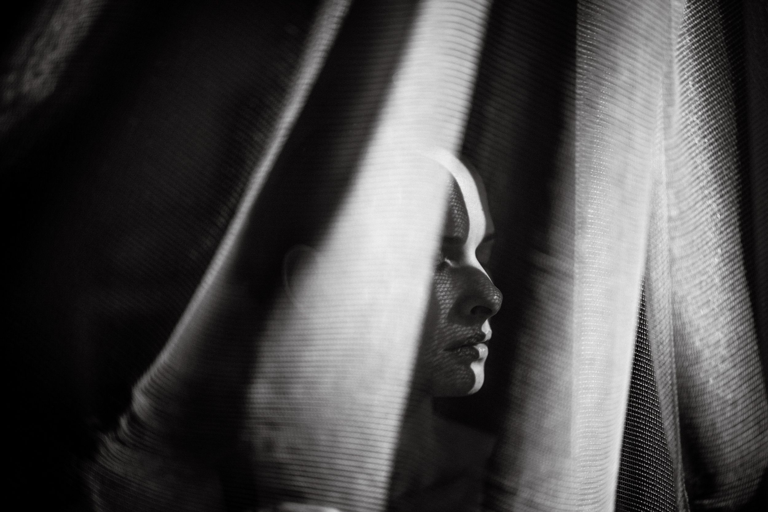 Black and white portrait of a person partially hidden behind flowing curtains, with soft light illuminating their face.