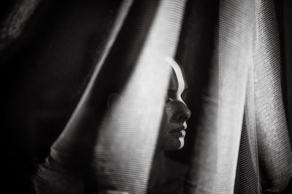 Black and white portrait of a person partially hidden behind flowing curtains, with soft light illuminating their face.