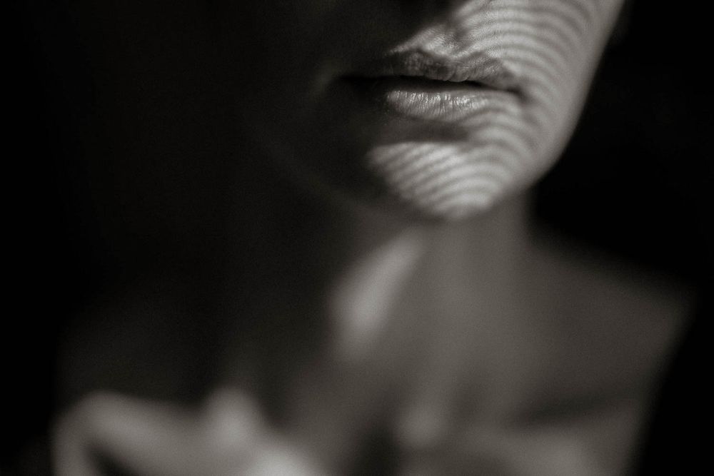 Black and white close-up portrait with dramatic striped shadow patterns across a woman's lips and neck.