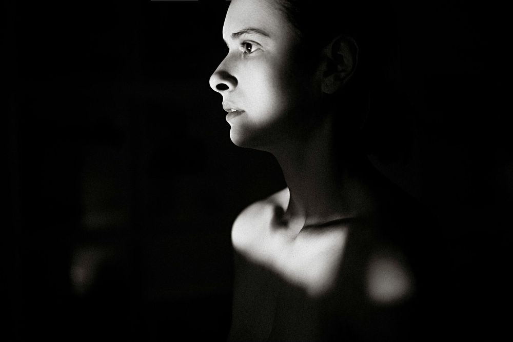 Black and white profile portrait of a young woman with dramatic side lighting against a dark background.