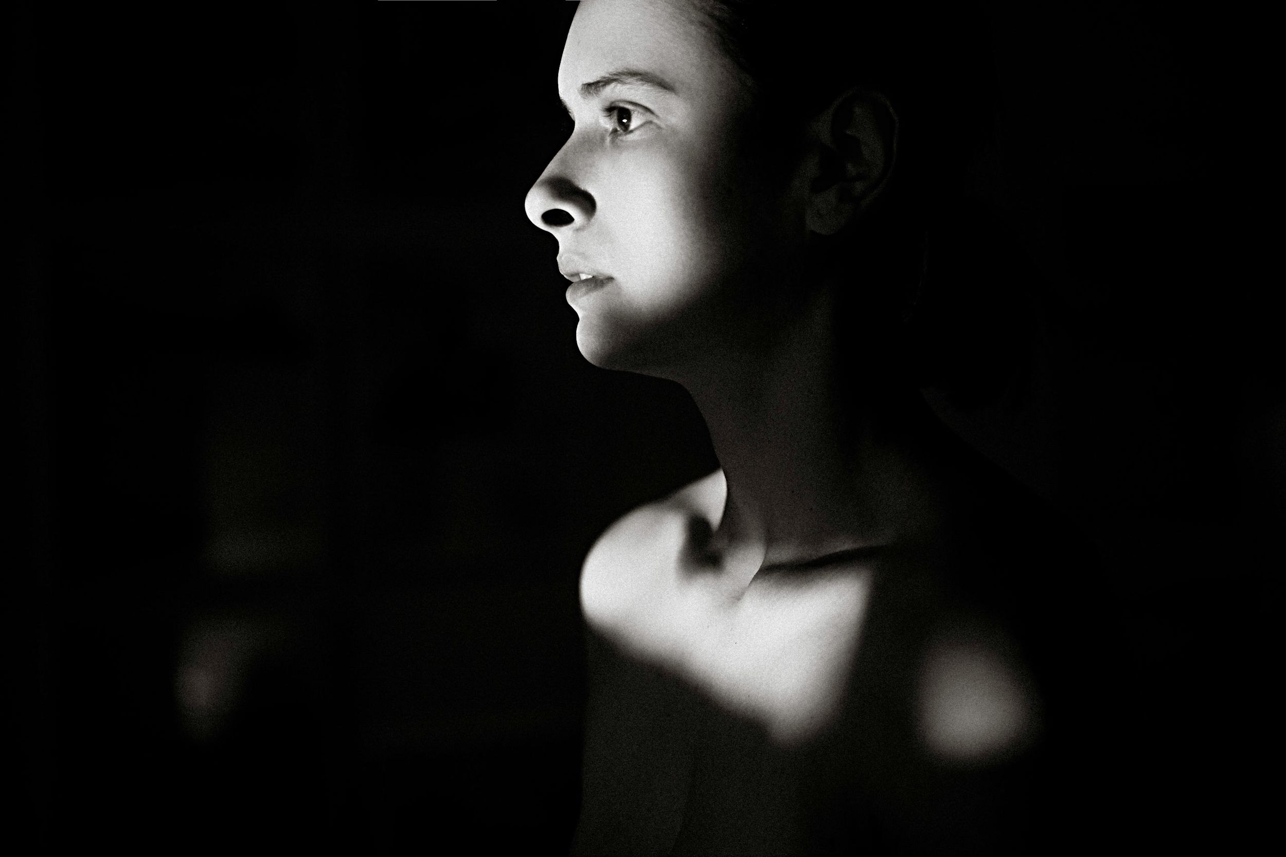 Black and white profile portrait of a young woman with dramatic side lighting against a dark background.
