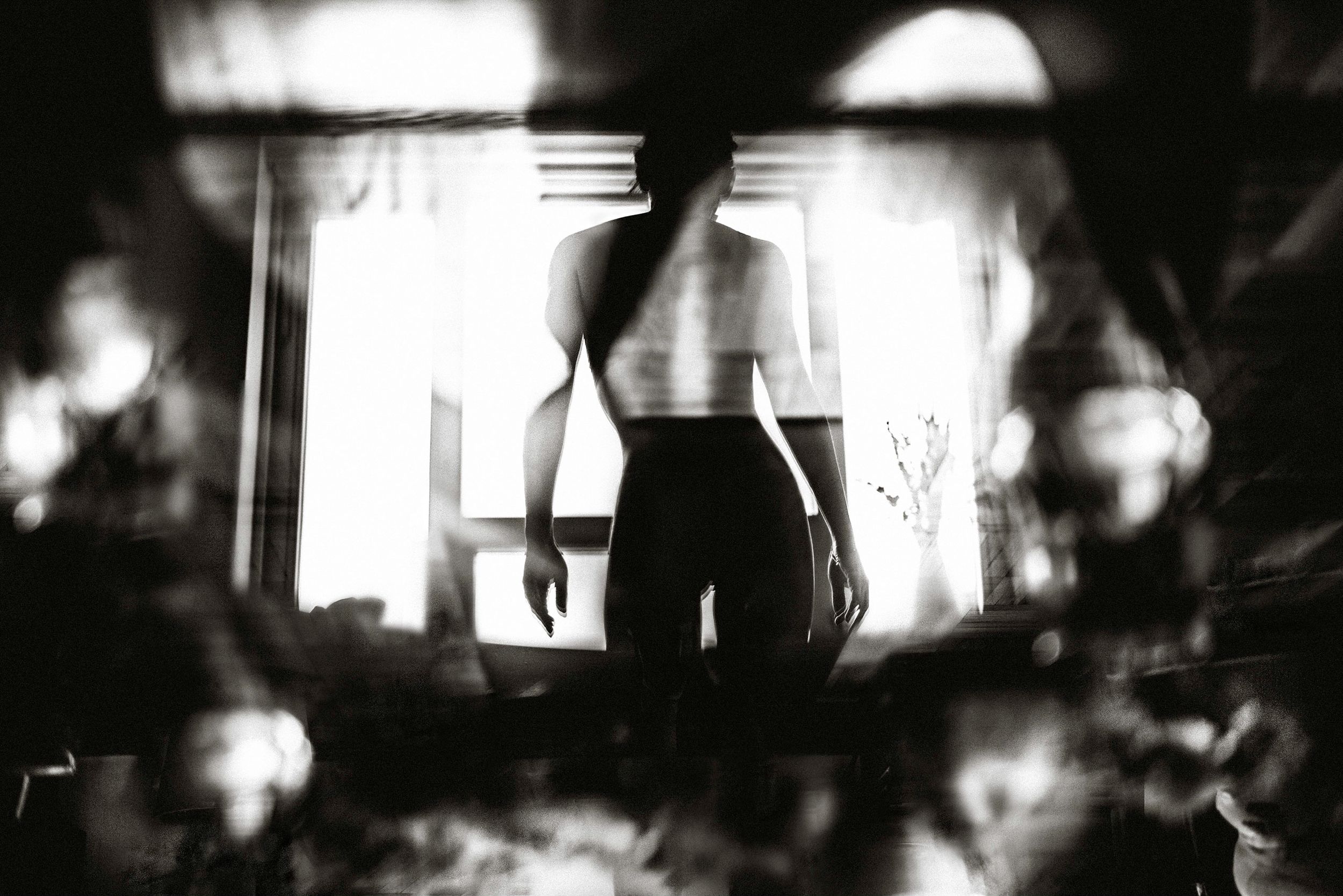 Black and white silhouette of a woman standing by a bright window in a dark, moody interior setting.