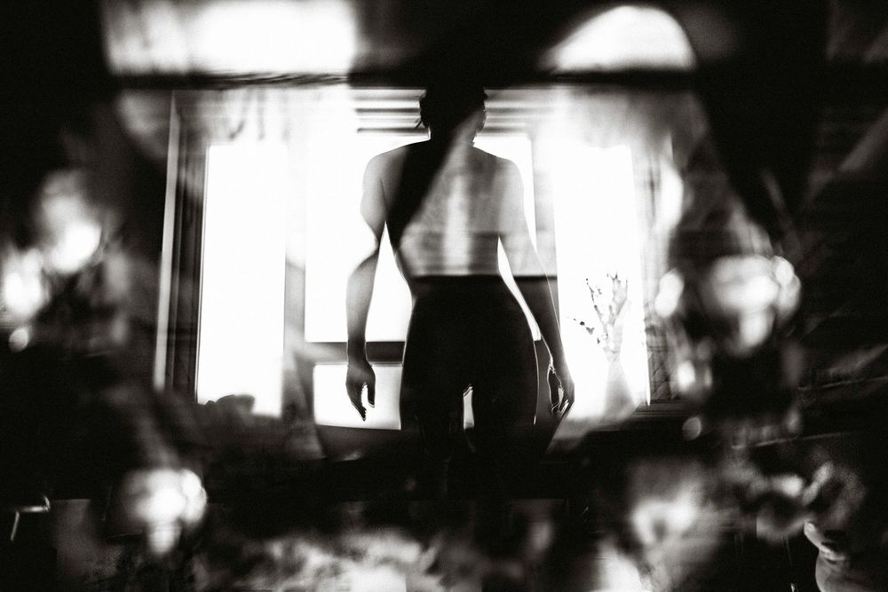 Black and white silhouette of a woman standing by a bright window in a dark, moody interior setting.