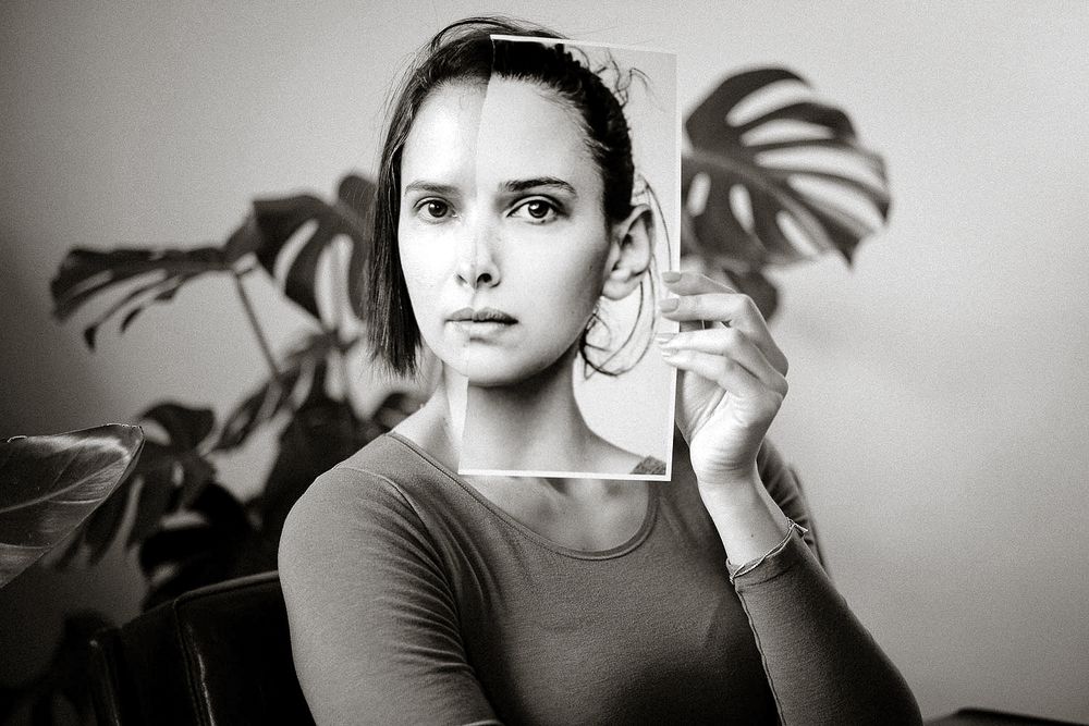 Black and white portrait of a woman holding a photo in front of her face beside a monstera plant.