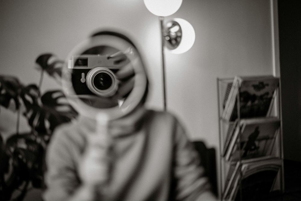 Black and white mirror selfie with a vintage film camera obscuring the face, indoor plants and lamp visible in background.
